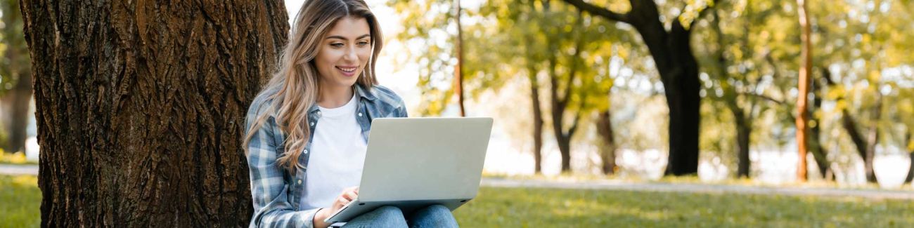 A female student sits quietly working on her online courses with her laptop. She is outside on a grassy, well landscaped lawn with her back against the rough bark of a mature evergreen tree.